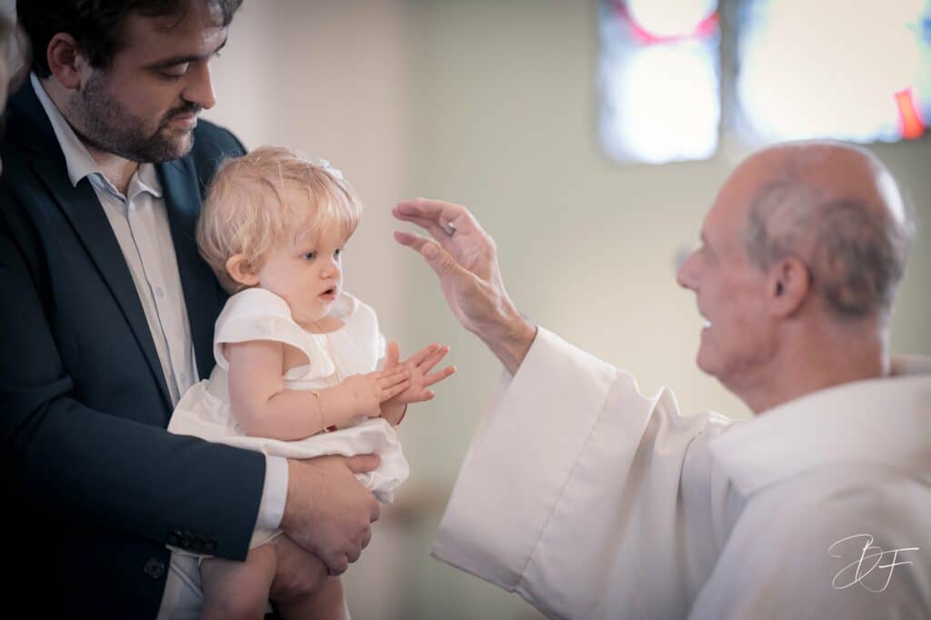 Le prêtre tend la main vers le front d'une petite fille en robe blanche, vitraux en arrière-plan, par BFO Photographie