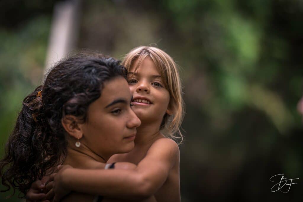 Adolescente serrant une petite fille blonde dans ses bras, joue contre joue, sur fond de verdure, par BFO Photographie