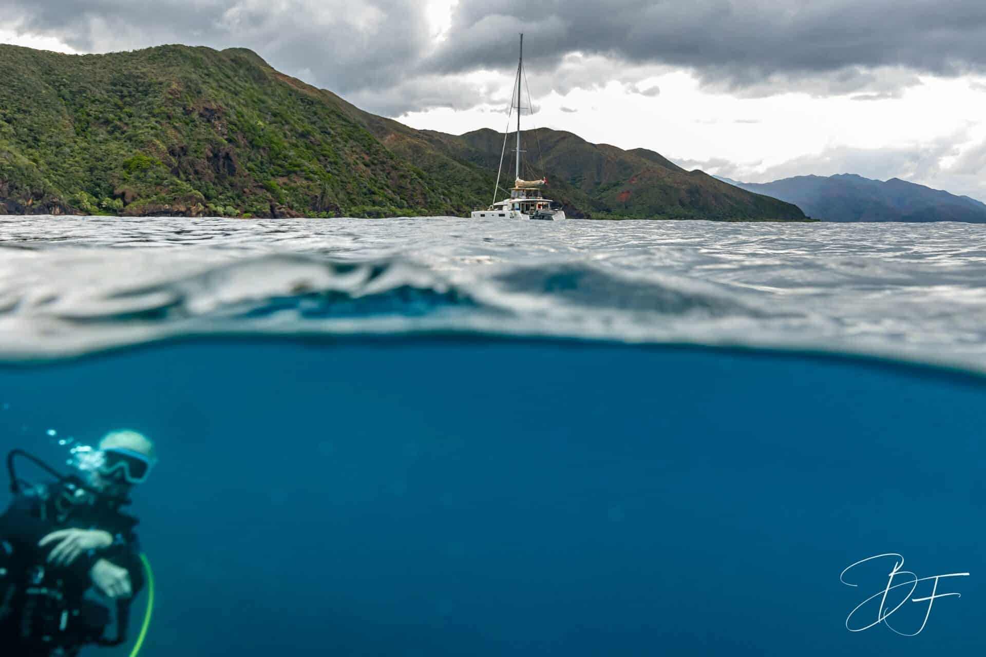Photo demi-sous-marine : plongeur sous la surface, catamaran et montagnes de la Côte Oubliée au-dessus, par BFO Photographie
