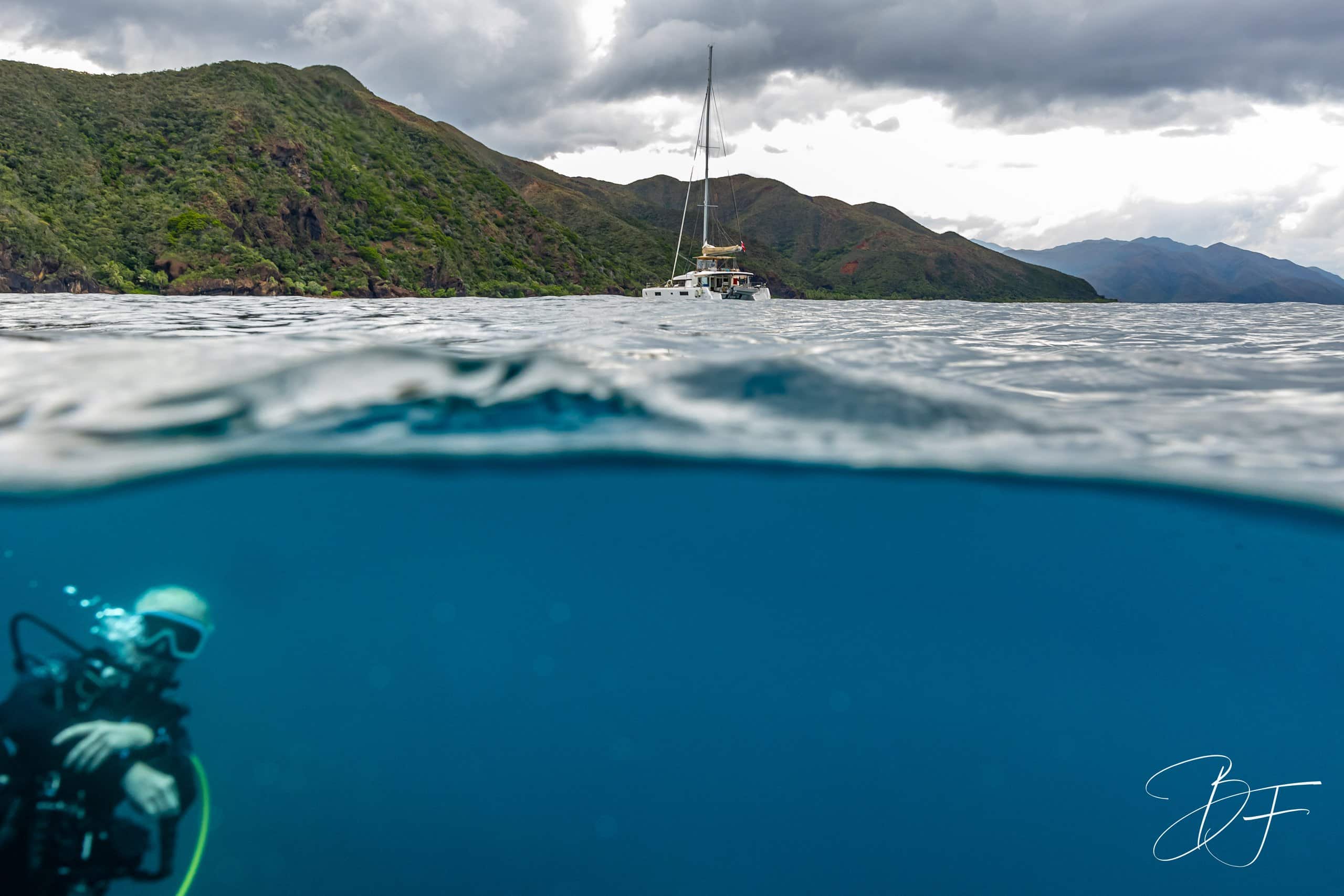 Over-under photo on the Côte Oubliée: a diver below the surface, a catamaran and wild mountains, by BFO Photographie
