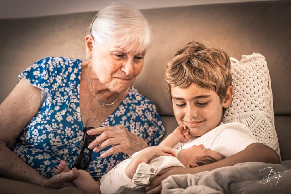 Grand-mère et jeune garçon souriant penchés sur un nouveau-né endormi, lumière chaude, par BFO Photographie
