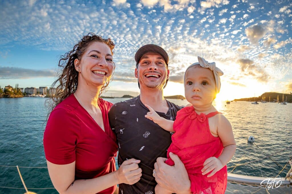 Parents et leur petite fille en rouge sur un bateau au coucher du soleil, baie de Nouméa en fond, par BFO Photographie