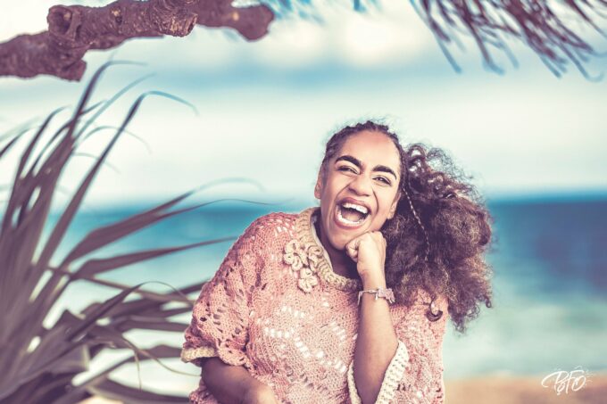 Esther éclate de rire, le poing sous le menton, cheveux au vent, au bord de la mer avec des feuilles de pandanus au premier plan